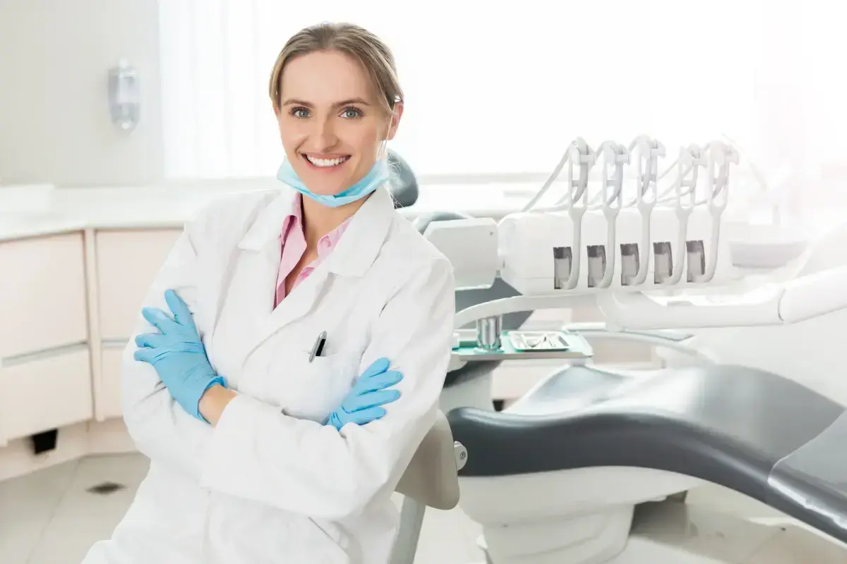 Female dentist in white coat and blue gloves smiling in modern dental office with treatment chair.